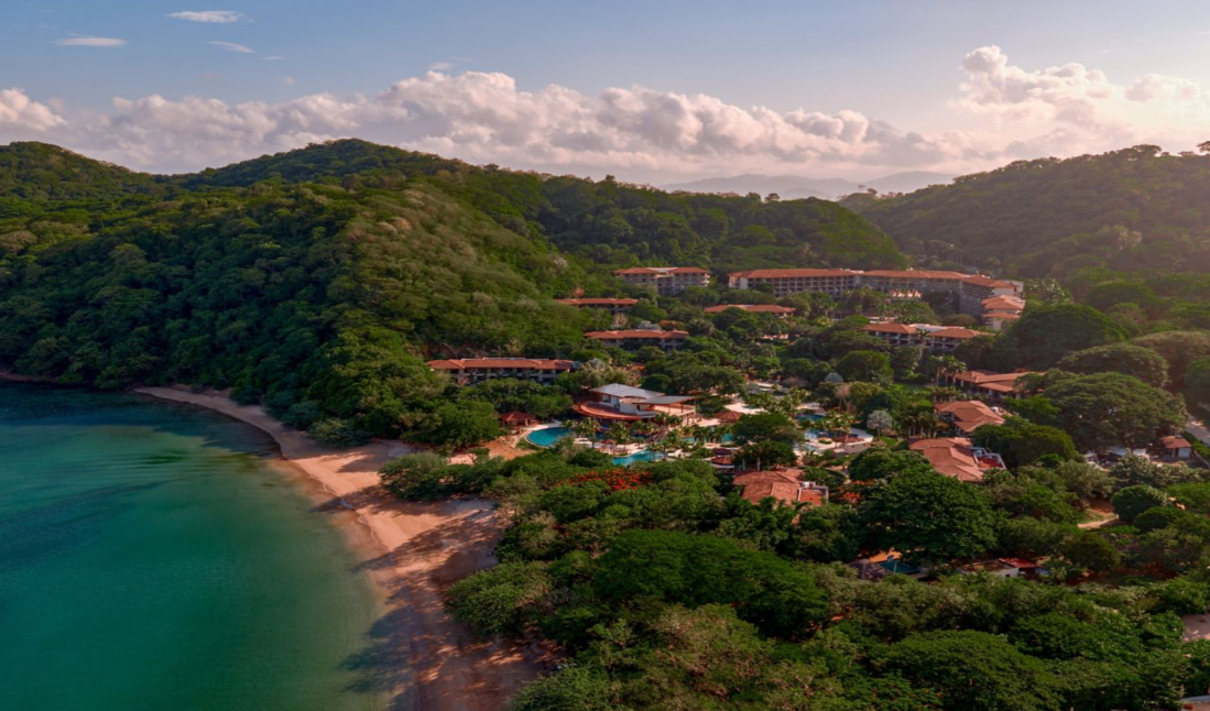 Aerial view of a beachfront resort surrounded by green hills beside a turquoise bay